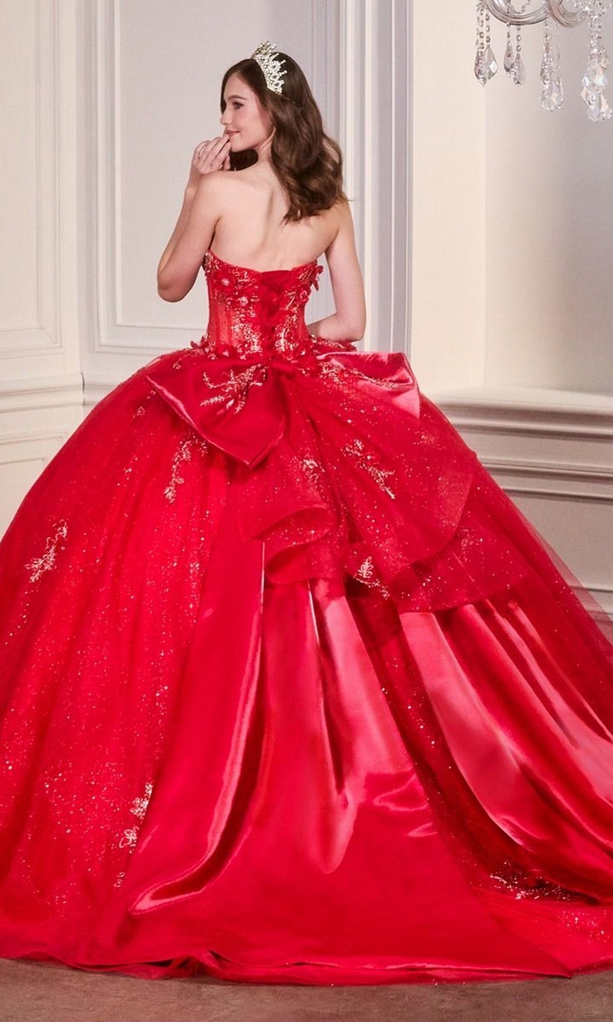 A woman stands indoors, partly turned away and touching her lips, wearing Ladivine’s Quinceanera Dress 15726—a sparkling strapless red ball gown with glitter tulle and a large back bow.